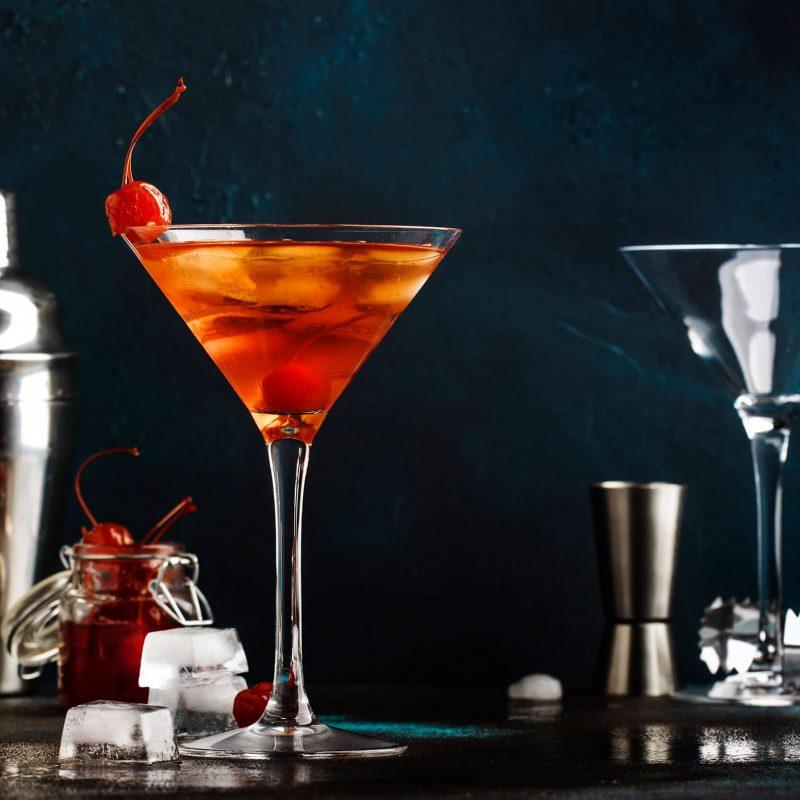 Sobre una mesada se ve una coctelera, un medidor, hielo, una copa de martini vacía y una copa de martini con un Mahattan, que es un cóctel anaranjado con hielo y una cereza en su interior.